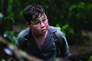 "A young boy with glasses, wearing a gray shirt and suspenders, looks upward with a concerned expression in a dense, green forest setting. His hair and face appear wet or sweaty, suggesting exertion or stress."